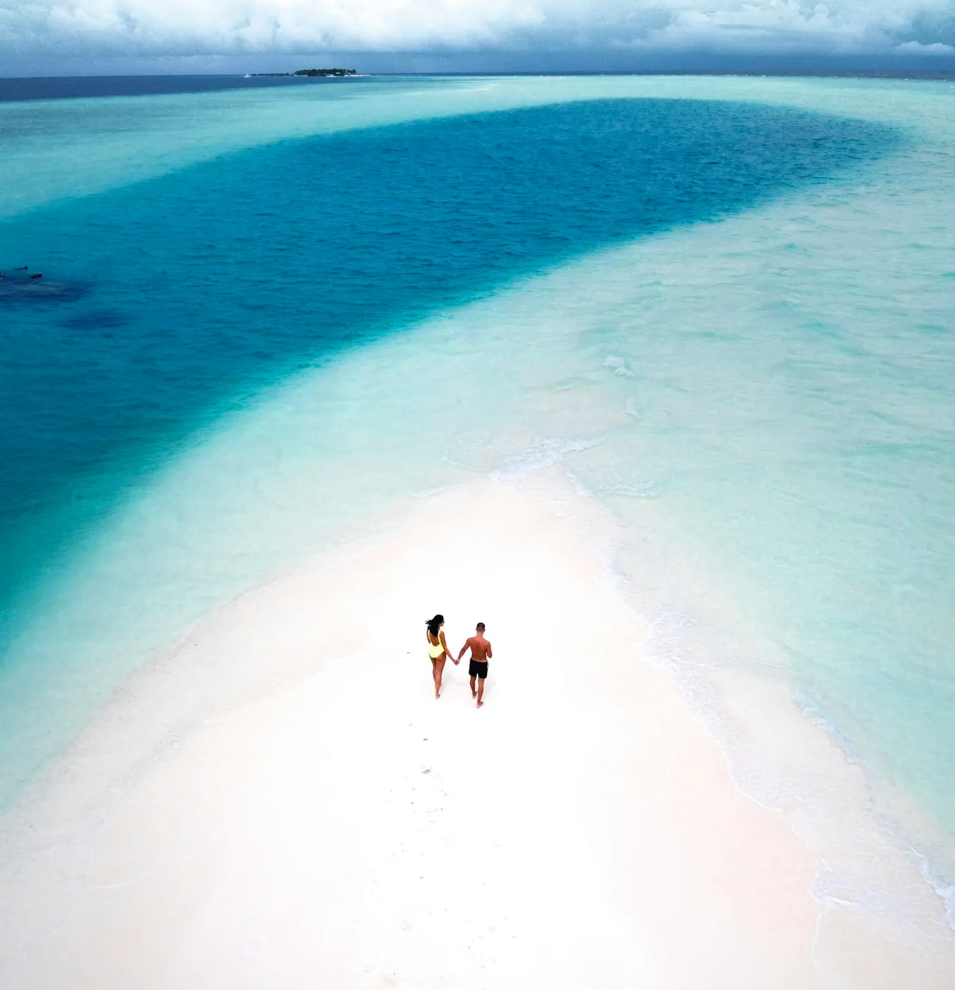 Two figures on a pale sandbar surrounded by shallow blue water